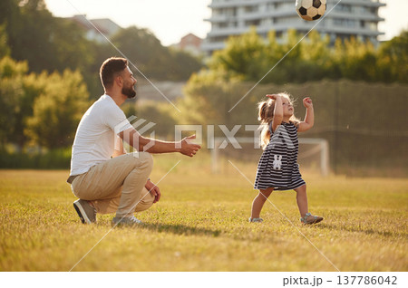 Throwing soccer ball high up. Father with his little daughter are on the field Throwing soccer ball high up. Father with his little daughter are on the field 137786042