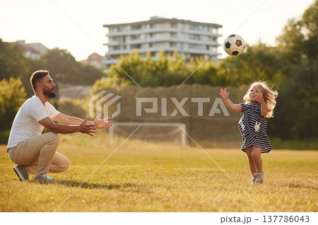 Throwing soccer ball high up. Father with his little daughter are on the field Throwing soccer ball high up. Father with his little daughter are on the field 137786043