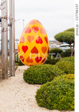 A big yellow Easter egg covered in red heart shapes is placed in a green garden near small bushes. It is daytime and shoppers can be seen in the background of the scene A big yellow Easter egg covered in red heart shapes is placed in a green garden near small bushes. It is daytime and shoppers can be seen in the background of the scene 137786197