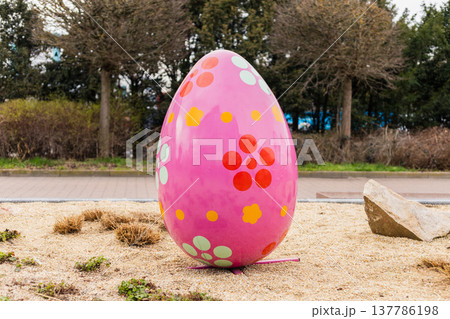A large pink egg with colorful flower patterns is placed on a sandy area in the garden. It is part of an Easter decoration setup. The setting includes trees and shrubs A large pink egg with colorful flower patterns is placed on a sandy area in the garden. It is part of an Easter decoration setup. The setting includes trees and shrubs 137786198