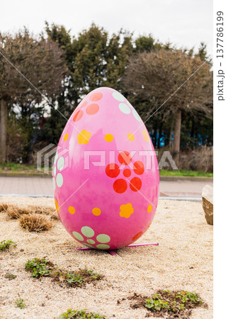 A large pink egg with colorful flower patterns is placed on a sandy area in the garden. It is part of an Easter decoration setup. The setting includes trees and shrubs A large pink egg with colorful flower patterns is placed on a sandy area in the garden. It is part of an Easter decoration setup. The setting includes trees and shrubs 137786199