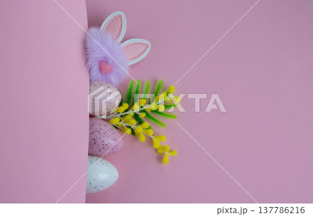 On a pink background, a fluffy rabbit head with ears and a pink nose is peeking out of a pocket, along with speckled eggs and artificial mimosa branches. Easter concept On a pink background, a fluffy rabbit head with ears and a pink nose is peeking out of a pocket, along with speckled eggs and artificial mimosa branches. Easter concept 137786216