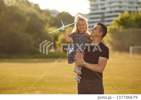 Man is holding girl high, she is with toy plane. Father with his little daughter are on the field 137786679