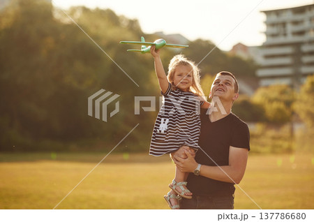 Man is holding girl high, she is with toy plane. Father with his little daughter are on the field 137786680