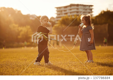 Conversation by using tin can telephone. Two kids are having fun on the summer field Conversation by using tin can telephone. Two kids are having fun on the summer field 137786884
