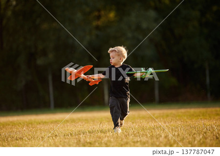 Running with two toy planes in hands. Boy is having fun on the field at daytime 137787047