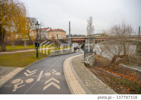 A bicycle path curves along the river in Prague. The scene shows a route marked for cyclists. Nearby, a bridge crosses the water. The sky appears gray, suggesting a cool day. 137787316