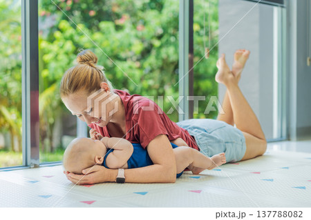 Mother and baby lying on a soft play mat at home, playing, talking and sharing a joyful moment together. Authentic family lifestyle, early childhood development and emotional bonding. Parenting and Mother and baby lying on a soft play mat at home, playing, talking and sharing a joyful moment together. Authentic family lifestyle, early childhood development and emotional bonding. Parenting and 137788082