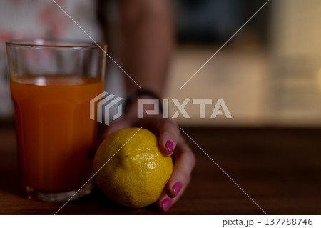 Intimate scene with lemon and polish, Textured lemon peel held next to glass on table surface, Detailed view of lemon peel and hand with nail polish beside glass on table 137788746