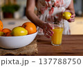 Woman holding lemon over glass, floral dress and wooden surface, bowl of citrus in foreground, fresh juice preparation, vitaminC wellness moment, influencerstyle natural light 137788750