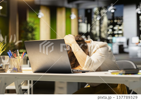 Businesswoman struggling to stay awake after an intense workload, lounges across desk at midnight. Fatigue, mental haze and workplace challenges have left her drained and restless. 137789329