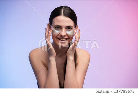 Young woman smiling while applying under-eye patches, showcasing a beauty routine for self-care, skin pampering, and promoting a concept of health and youthfulness Young woman smiling while applying under-eye patches, showcasing a beauty routine for self-care, skin pampering, and promoting a concept of health and youthfulness 137789675