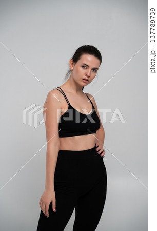 Young adult woman standing confidently in a studio, wearing black sports bra and leggings, promoting concepts of fitness, health, vitality, and an active lifestyle Young adult woman standing confidently in a studio, wearing black sports bra and leggings, promoting concepts of fitness, health, vitality, and an active lifestyle 137789739