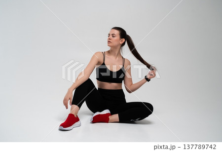 Fitness woman sitting on white background, wearing black sportswear and red sneakers, pulling her long ponytail, demonstrating a healthy and active lifestyle Fitness woman sitting on white background, wearing black sportswear and red sneakers, pulling her long ponytail, demonstrating a healthy and active lifestyle 137789742