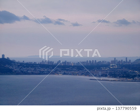 Serene Shoreline Beneath Cloudy Urban Horizon, Calm Waters Meet Foggy City Skyline In Gentle Mist 137790559