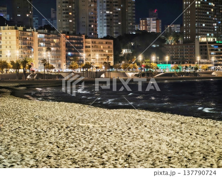 Textured Beach Front Leading To Glowing Cityscape With Ocean Waves And Silhouettes Of Towers 137790724
