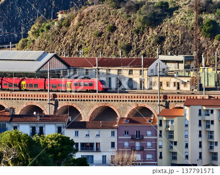 Vibrant Depiction Of Red Train Journeying Through Terraced Cityscape With Stone Bridge And Sunset Glow 137791571