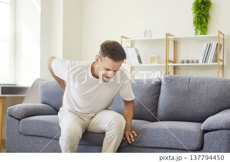 Sick man with lower back pain sitting on sofa at home, suffering from backache Sick man with lower back pain sitting on sofa at home, suffering from backache 137794450