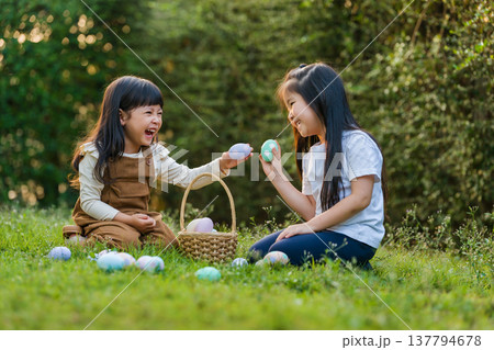 two happy child girl playing together and hunting for Easter eggs into basket on green grass in garden two happy child girl playing together and hunting for Easter eggs into basket on green grass in garden 137794678