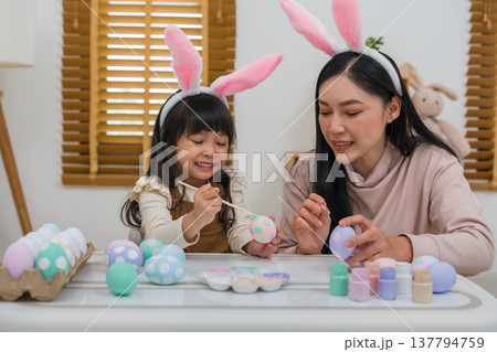 mother and preschool child girl wearing bunny ears painting and decorating egg together at home on easter day. happy family holiday activity concept mother and preschool child girl wearing bunny ears painting and decorating egg together at home on easter day. happy family holiday activity concept 137794759