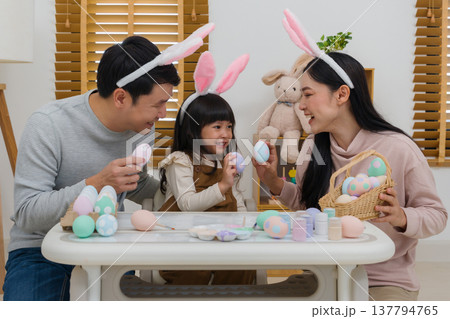 father, mother and daughter wearing bunny ears holding colorful egg together at home on easter day. happy family holiday activity concept father, mother and daughter wearing bunny ears holding colorful egg together at home on easter day. happy family holiday activity concept 137794765