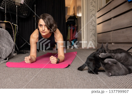 A woman performs a forearm plank on a pink mat at home while kittens play in the foreground. 137799907