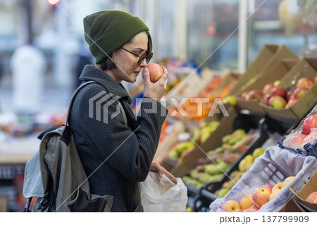 Side view of a woman in sunglasses and a beanie smelling a red apple while shopping at an outdoor fruit stand with a reusable bag. Side view of a woman in sunglasses and a beanie smelling a red apple while shopping at an outdoor fruit stand with a reusable bag. 137799909