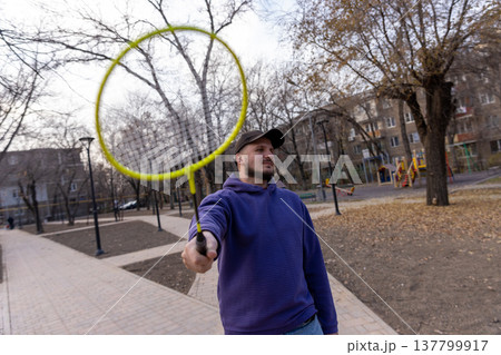 A playful point-of-view shot of a man holding a yellow badminton racket directly at the lens in an autumn park. 137799917