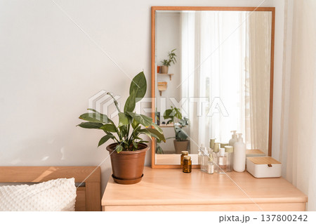 Wooden vanity chest of drawers holding a potted plant, various skincare bottles, and a mirror reflecting a bright window. Vanity dressing table with plant and skincare products Wooden vanity chest of drawers holding a potted plant, various skincare bottles, and a mirror reflecting a bright window. Vanity dressing table with plant and skincare products 137800242