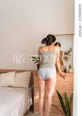 Woman standing in bedroom, looking at her body reflection in the dresser mirror, practicing self-care and body positivity. Adult woman admiring body reflecting in mirror at home Woman standing in bedroom, looking at her body reflection in the dresser mirror, practicing self-care and body positivity. Adult woman admiring body reflecting in mirror at home 137800244