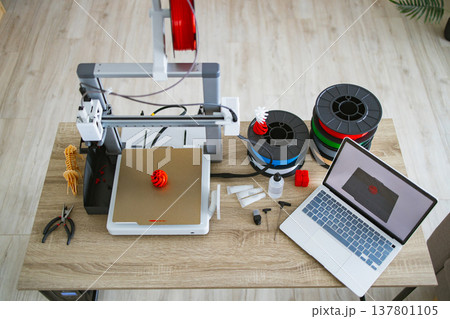A 3D printer is set up on a wooden desk with spools of filament, tools, and a laptop displaying a 3D model 137801105