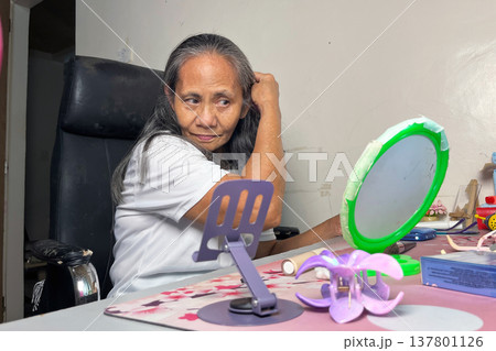 Senior woman sitting at desk and adjusting her hair in front of mirror, elderly lady doing self-care routine in her room Senior woman sitting at desk and adjusting her hair in front of mirror, elderly lady doing self-care routine in her room 137801126