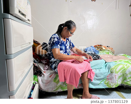 Senior woman sitting on bed and folding clothes in cozy bedroom, elderly lady organizing laundry at home, domestic lifestyle portrait Senior woman sitting on bed and folding clothes in cozy bedroom, elderly lady organizing laundry at home, domestic lifestyle portrait 137801129