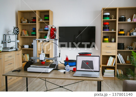 A modern 3D printing workstation is set up on a desk with multiple printers, filament spools, and a laptop displaying a design A modern 3D printing workstation is set up on a desk with multiple printers, filament spools, and a laptop displaying a design 137801140