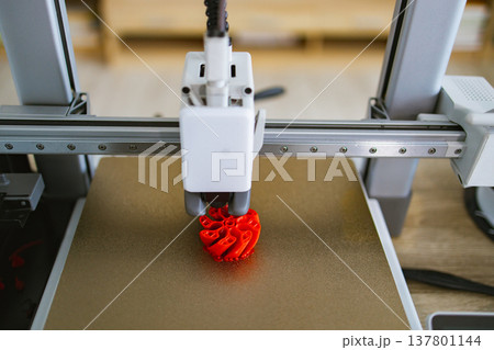 A close-up view of a 3D printer's nozzle depositing molten plastic to build a complex red object layer by layer on a gold build plate 137801144
