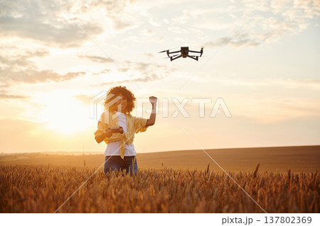 Modern black drone is up. Beautiful woman is on the wheat agricultural field Modern black drone is up. Beautiful woman is on the wheat agricultural field 137802369