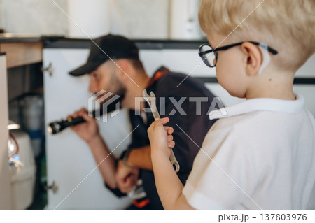 Little boy is helping, holding tool for plumber that is working on the kitchen Little boy is helping, holding tool for plumber that is working on the kitchen 137803976