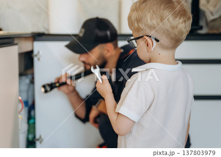 Little boy is helping, holding tool for plumber that is working on the kitchen Little boy is helping, holding tool for plumber that is working on the kitchen 137803979
