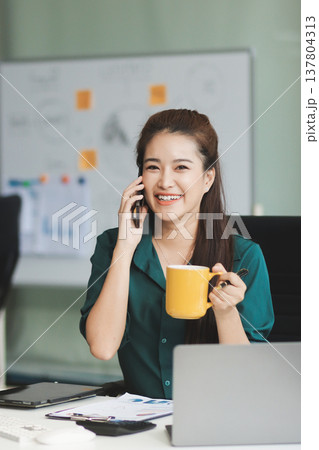 Determined asian businesswoman works late in a modern office, confidently analyzing data on her laptop, embodying success and professionalism with a smile 137804313