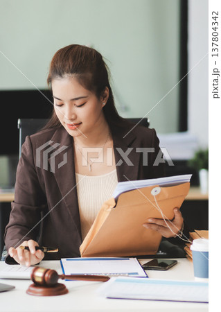 Confident Lawyer Working at Desk: A young Asian female lawyer confidently works on her laptop, surrounded by legal symbols. She exudes professionalism and competence in her modern office setting. Confident Lawyer Working at Desk: A young Asian female lawyer confidently works on her laptop, surrounded by legal symbols. She exudes professionalism and competence in her modern office setting. 137804342