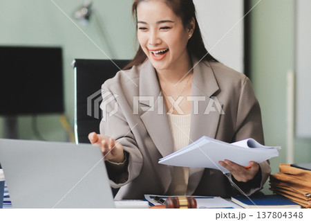 Confident Lawyer Working at Desk: A young Asian female lawyer confidently works on her laptop, surrounded by legal symbols. She exudes professionalism and competence in her modern office setting. 137804348
