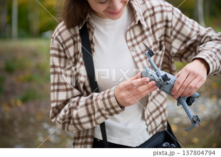A young woman carefully inspects her drone while standing in a scenic area filled with fall colors. The moment captures her passion for technology and exploration in nature. A young woman carefully inspects her drone while standing in a scenic area filled with fall colors. The moment captures her passion for technology and exploration in nature. 137804794