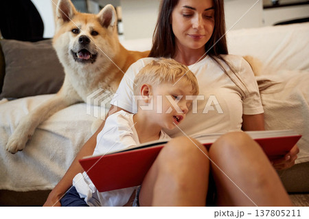 Leaning on the bed, resting, reading. Mother with son and Shibu Inu dog are at home Leaning on the bed, resting, reading. Mother with son and Shibu Inu dog are at home 137805211