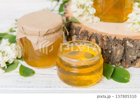 Sweet honey jar surrounded spring acacia blossoms. Honey flows from a spoon in a jar. jars of clear fresh acacia honey on wooden background Sweet honey jar surrounded spring acacia blossoms. Honey flows from a spoon in a jar. jars of clear fresh acacia honey on wooden background 137805310