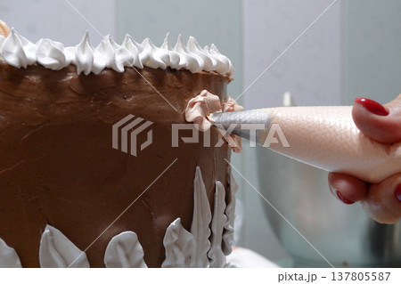 A woman pastry chef with a pastry bag in her hands squeezes cream on a cake in a home kitchen. Professional chef at work on bakery kitchen. culinary, baking and cooking food concept, home cooking A woman pastry chef with a pastry bag in her hands squeezes cream on a cake in a home kitchen. Professional chef at work on bakery kitchen. culinary, baking and cooking food concept, home cooking 137805587