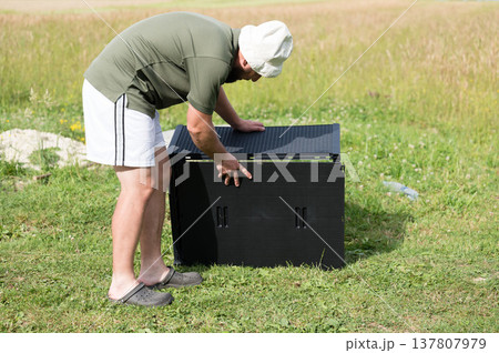 Man assembling a black plastic storage unit outdoors on a grassy area. Man assembling a black plastic storage unit outdoors on a grassy area. 137807979