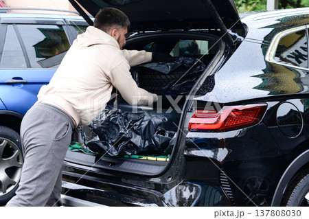 Man loading black bags into open trunk of black SUV in parking lot on a rainy day in the afternoon Man loading black bags into open trunk of black SUV in parking lot on a rainy day in the afternoon 137808030