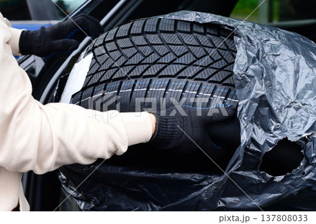 Person carries new tire in black plastic bag toward car in a parking lot during daytime in autumn season Person carries new tire in black plastic bag toward car in a parking lot during daytime in autumn season 137808033