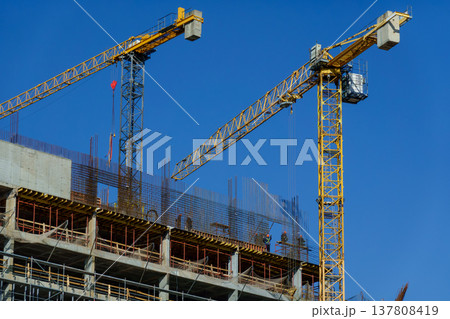 Construction site with tower cranes and workers assembling concrete high rise under clear blue sky Construction site with tower cranes and workers assembling concrete high rise under clear blue sky 137808419
