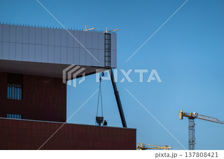 Construction crane lifting concrete bucket beside modern building under clear blue sky 137808421
